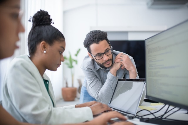 Duas mulheres e um homem em um ambiente de escritório. Uma das mulheres está concentrada em seu notebook enquanto o homem se inclina para observar a tela e conversar sobre o projeto, representando a colaboração técnica entre profissionais de áreas não-dev.